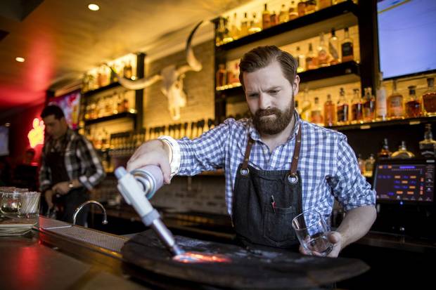 A bartender lights fire to a piece of wood so he can smoke a dish under a glass dome.