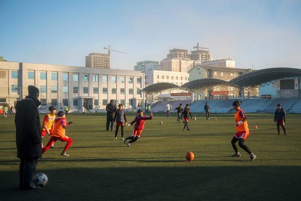 Children play an organized game of soccer after school. In this sports-mad society, pitches such as this one with artificial grass and bleachers aren’t an uncommon sight.