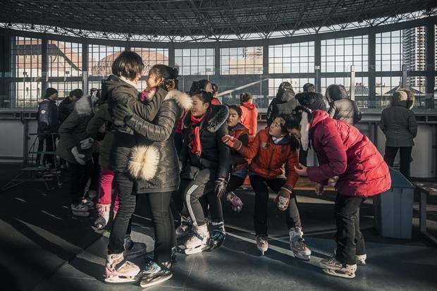 A gaggle of teenaged girls jostle for position and balance on a lace-up bench during a school outing to a skating rink. Though the ice surface itself is in dire need of a Zamboni, the students are good sports, and boys and girls stick to separate corners of the ice.