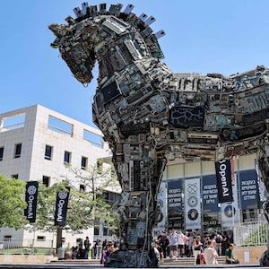 An art installation of a Trojan Horse comprised of e-waste appears on the Tel Aviv University campus while banners for OpenAI adorn a nearby building in 2023. A group of researchers left OpenAI and founded Anthropic in 2021 over concerns about the company’s resistance to guardrails to help regulate its AGI ambitions.