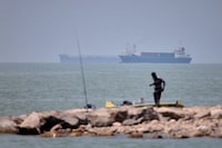 A man stands beside a fishing pole along the shore as cargo ships and commercial vessels are seen in the Strait of Hormuz off Bandar Abbas, Iran, Wednesday, June 17, 2026. (Amirhosein Khorgooi/ISNA via AP)