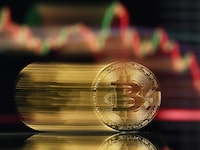 An illustration photograph taken on November 22, 2025, shows a gold plated souvenir Bitcoin coin reflected in a mirror and arranged for a photograph in front of a computer screen displaying the Bitcoin monthly price chart in a residential property in Guildford, south of London. The value of bitcoin has fallen sharply since reaching record heights last month, above $126,000 at the start of October. (Photo by Justin TALLIS / AFP via Getty Images)