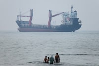 A group of people stands in shallow water as a cargo ship appears anchored in the Strait of Hormuz off Bandar Abbas, Iran, Tuesday, June 30, 2026. (Amirhosein Khorgooi/ISNA via AP)