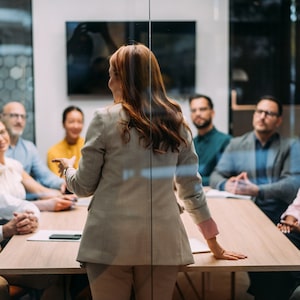Rear view shot of an unrecognizable businesswoman giving a presentation in the office board room. Group of business persons in business meeting. Creative business team on meeting in the office. The view is through glass.
